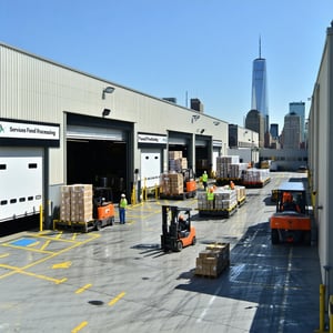 The image depicts a bustling loading dock area in an urban setting with a backdrop of the New York City skyline Various industrial vehicles including trucks and forklifts are seen actively loading and unloading goods Workers in safety gear maneuver a